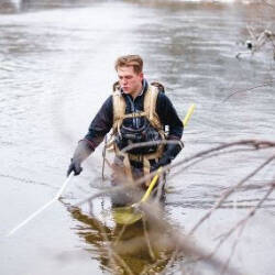 A person wades in a river, wearing a backpack and holding two poles, surrounded by bare trees.